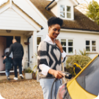 A woman attaches a charger to her electric vehicle as her family walks into their home. 